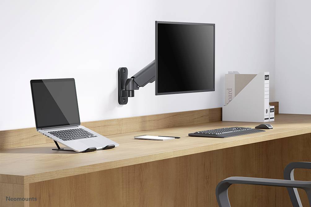 Minimalistický psací stůl s laptopem na stojanu, monitor uchycený na nástěnném držáku, klávesnice, myš a složky; moderní kancelářské vybavení.