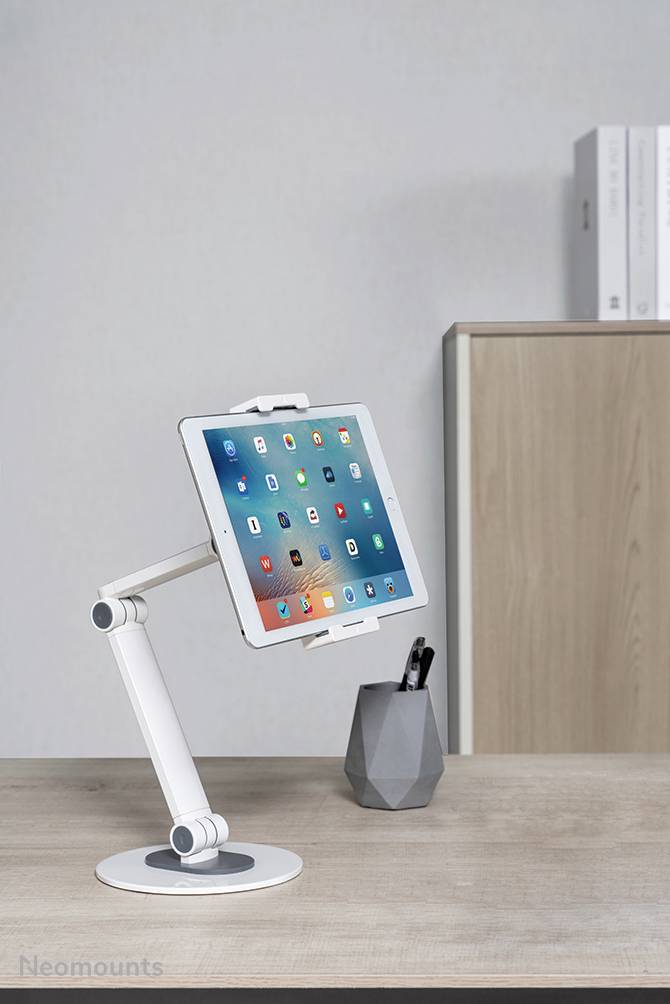 Tablet na flexibilním stolním stojanu, umístěný na psacím stole s držákem na tužky; v pozadí knihovna.