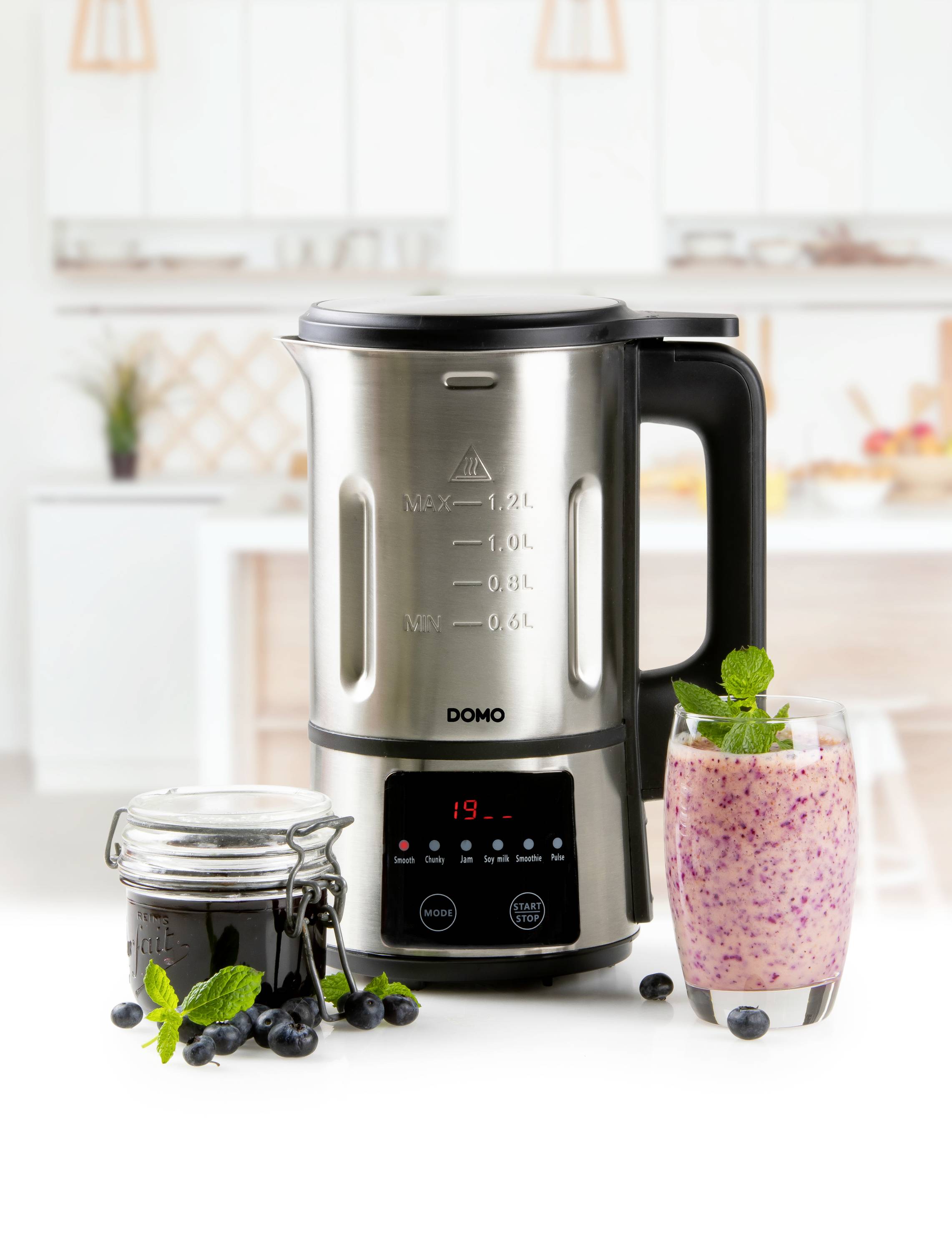 Na kuchyňské pracovní desce stojí mixér s sklenicí smoothie a sklenicí marmelády vedle něj. V pozadí je vidět kuchyň.