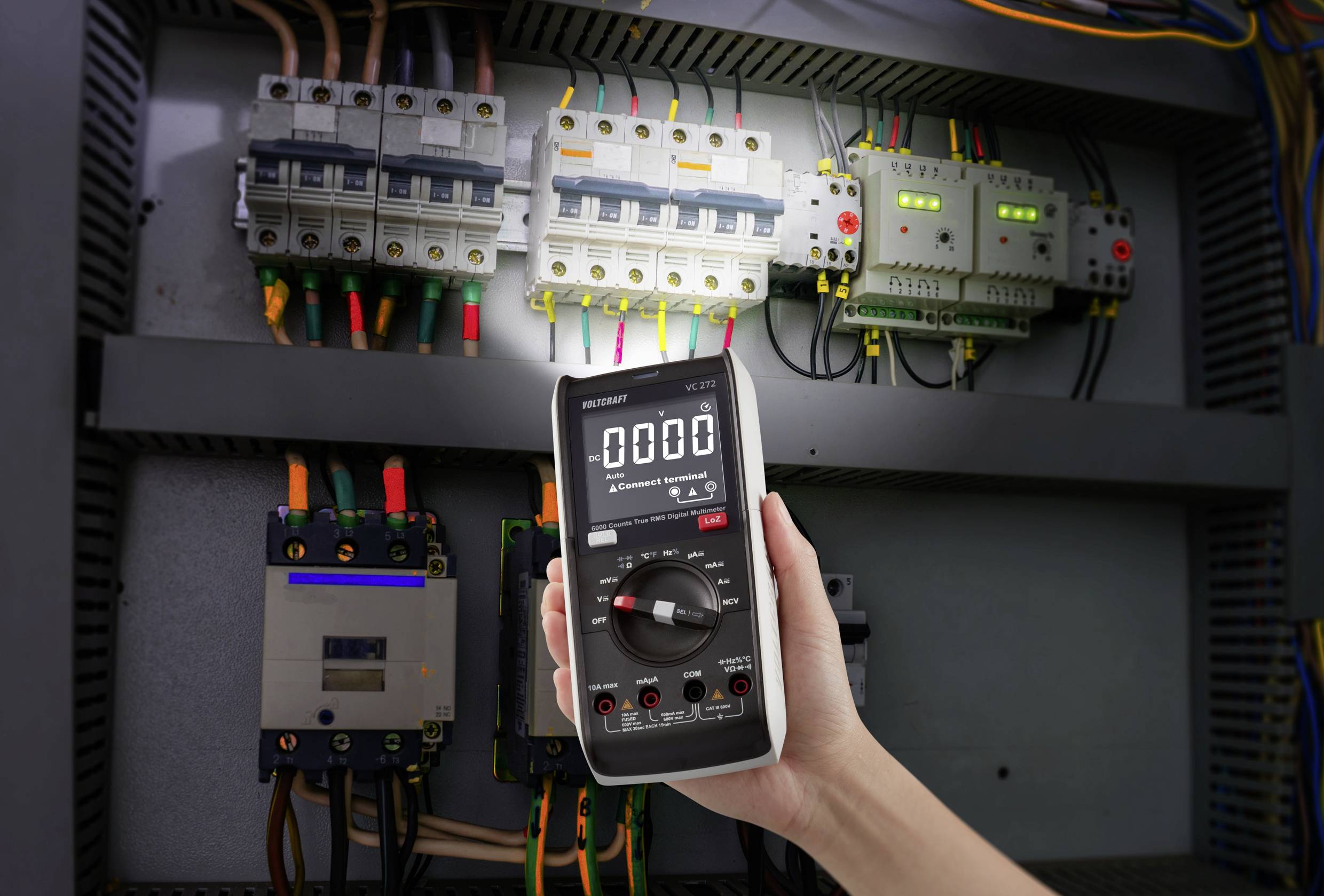 Jedna ruka drží digitální multimetr ukazující nulu před otevřenou elektrickou rozvodnou skříní s pojistkami a kabely.