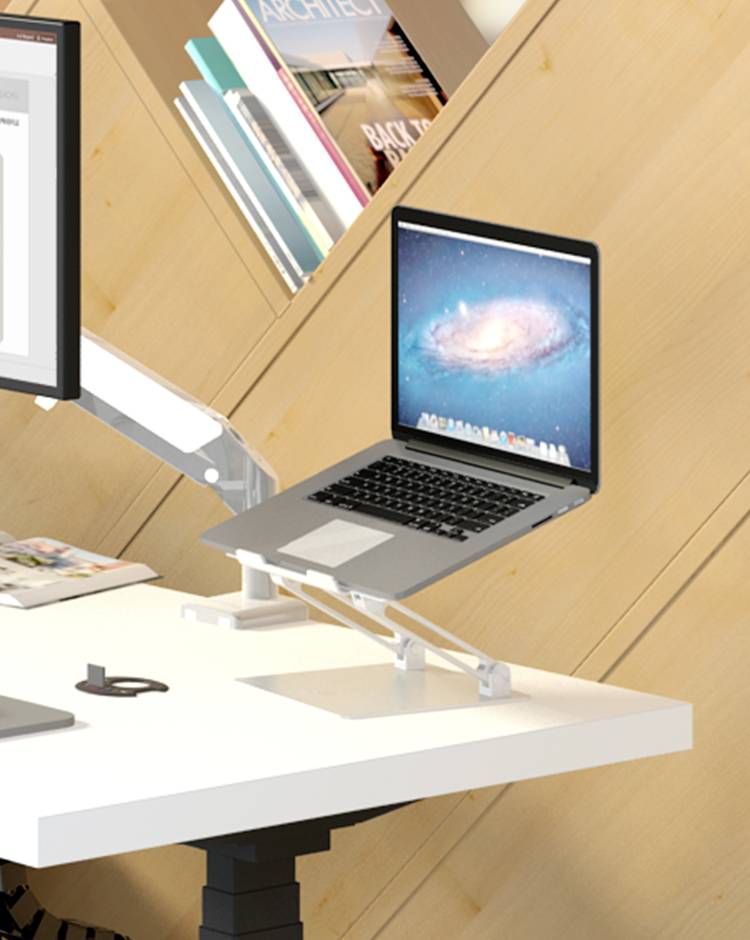 Notebook na nastavitelném stojanu vedle velkého monitoru na bílém psacím stole; v pozadí knihovny.