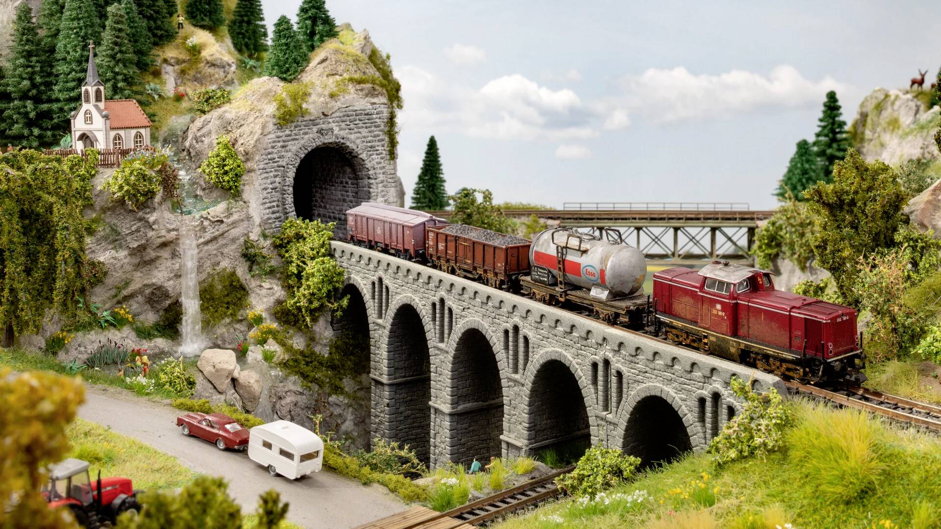 Model vláčku jede přes kamenný most, který vychází z tunelu. Na kopci stojí stromy a malý kostelík. V popředí jsou vidět automobil a obytný přívěs.