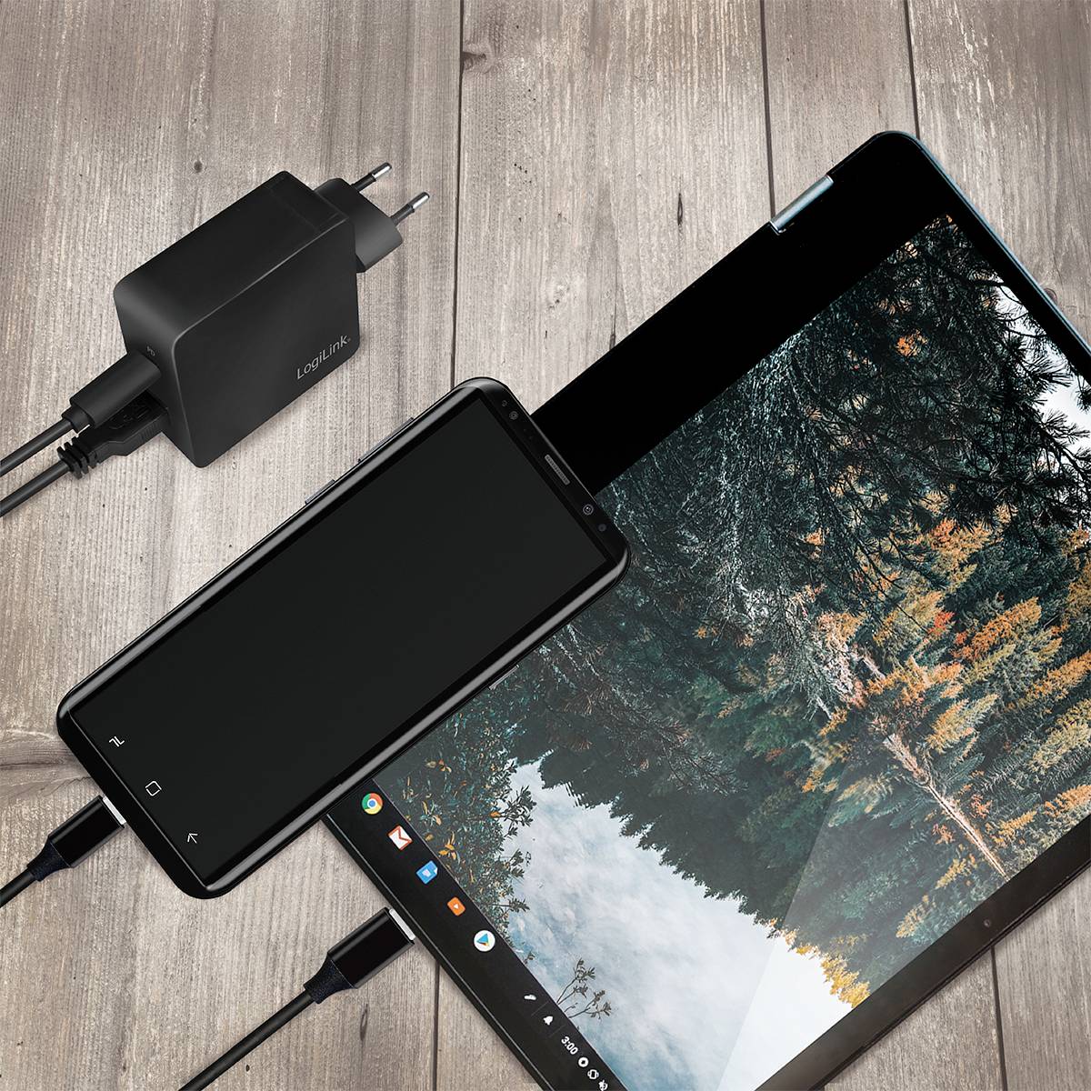 Mobilní telefon a tablet ležící na dřevěném stole jsou oba nabíjeny. Vedle nich leží nabíječka s evropskou zástrčkou.