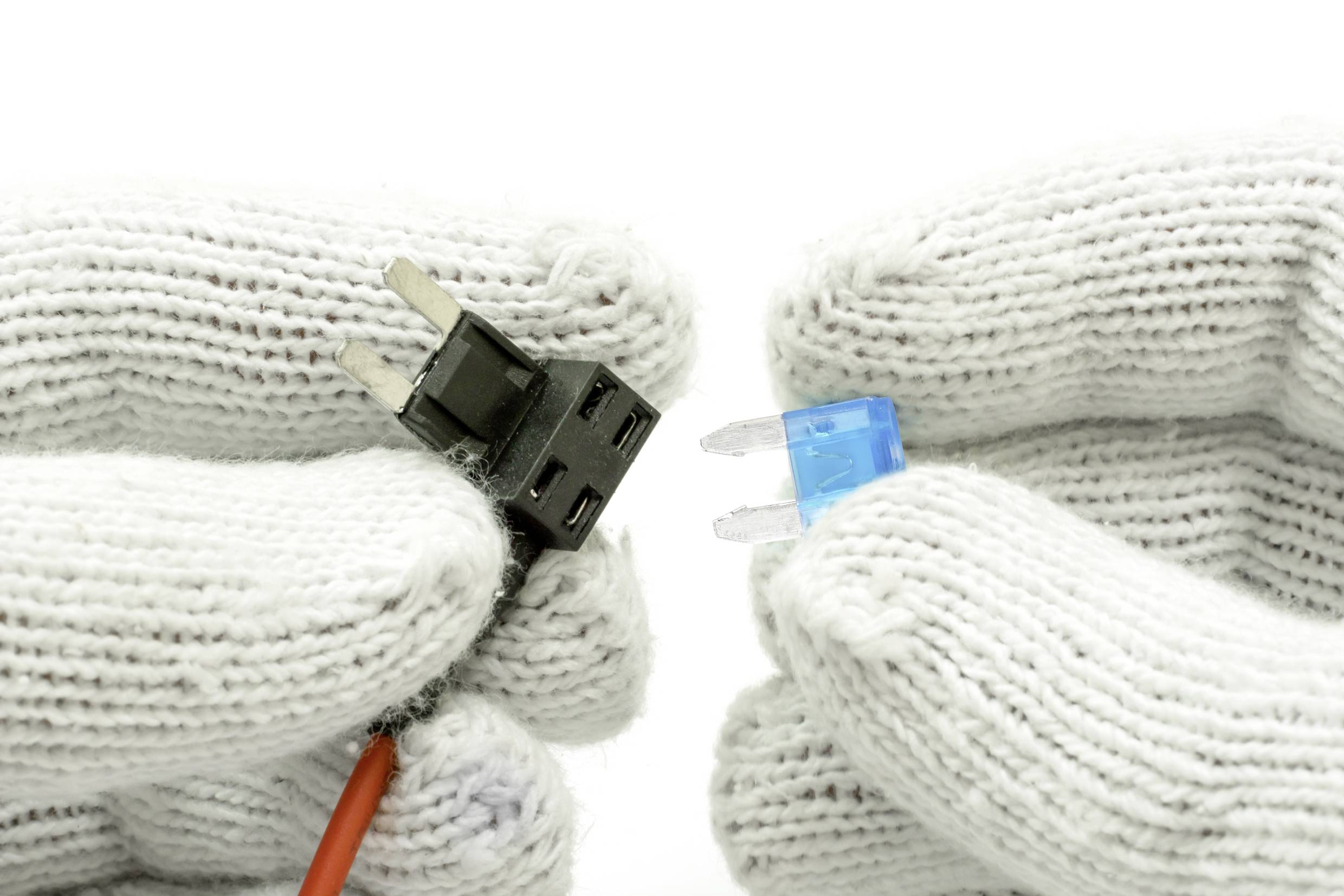 Dvě bílé rukavice drží elektrické komponenty: vlevo černá zástrčka s kabelem, vpravo modrá pojistka.