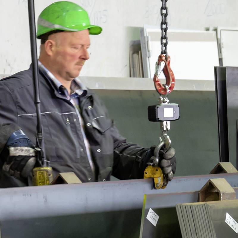 Dělník v zelené přilbě obsluhuje jeřáb v průmyslové hale. Jsou přemisťovány kovové desky. Jsou viditelné bezpečnostní boty a rukavice.
