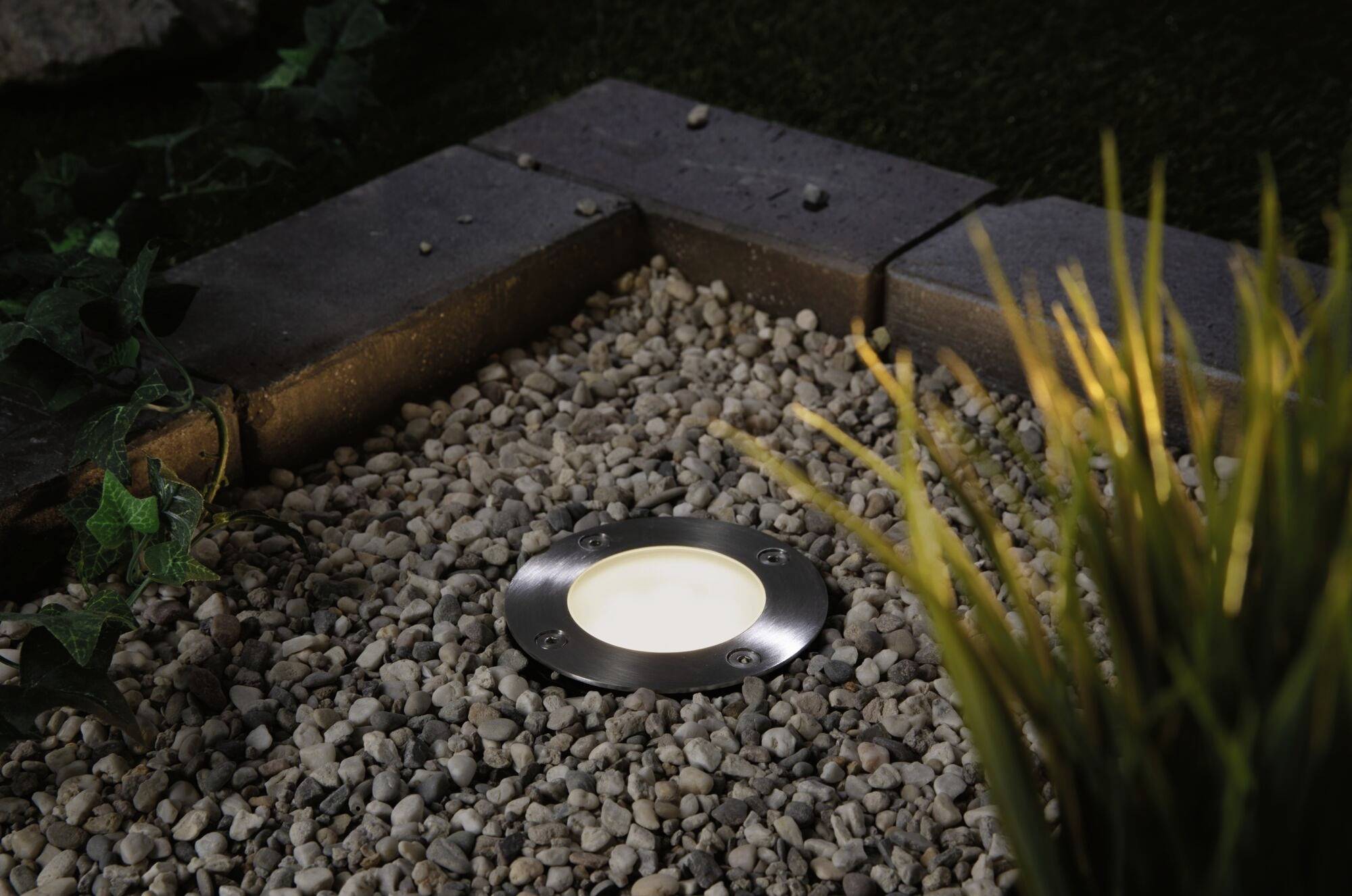 Zemní svítidlo v zahradní části pokryté štěrkem, obklopené rostlinami a obrubníky trávníku. Lampa svítí jemně.