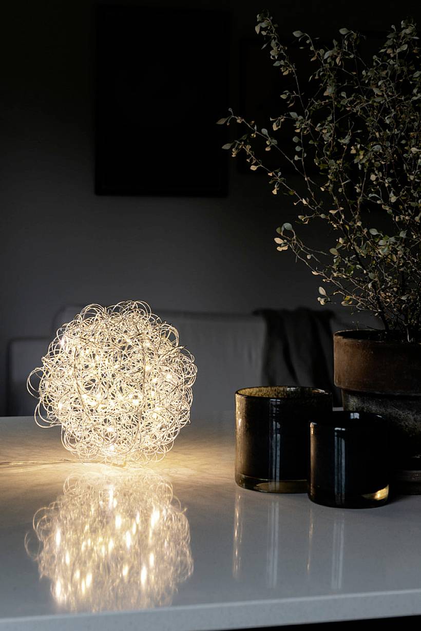 Osvětlená drátěná lampa ve tvaru koule stojí na stole vedle dvou tmavých svícnů a pokojové rostliny v tmavé místnosti.