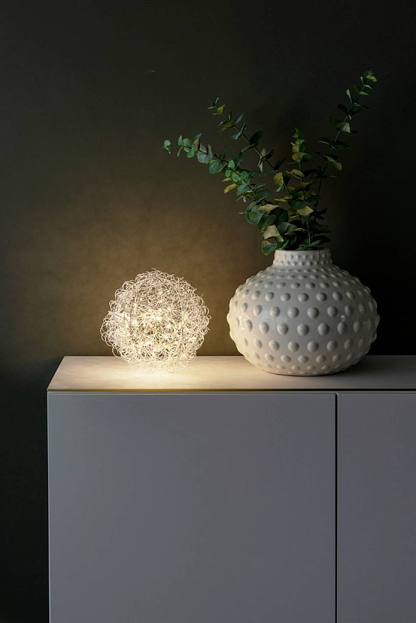 Stůl s dekorativní lampou a bílou vázu s rostlinami před tmavým pozadím. Lampa má kulovitý drátěný design.