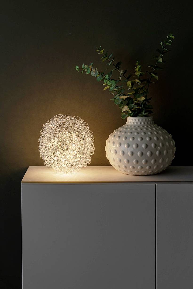Stolní lampa s designem drátěné mřížky jemně osvětluje prostor vedle bílé vázy s zeeleným větvemi umístěné na šedé skříňce.