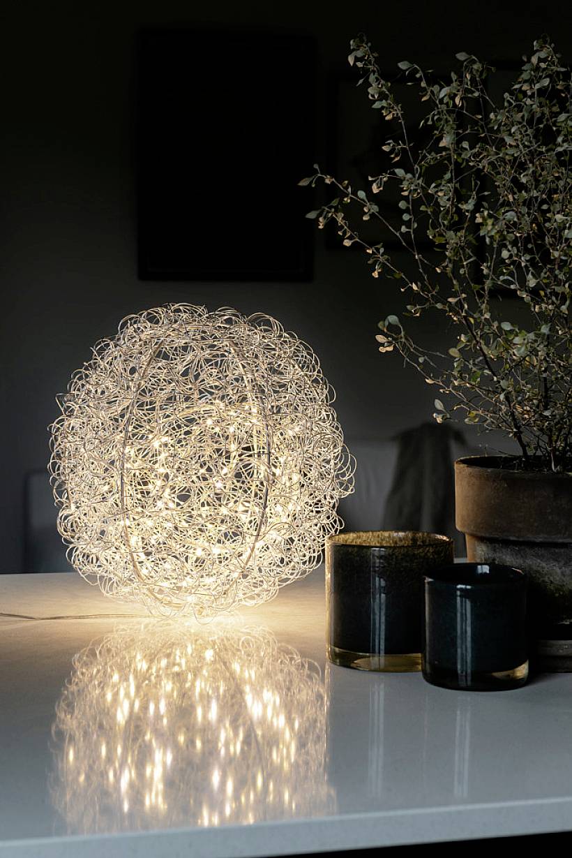 Zářivá, kulovitá stolní lampa z kroucené drátu stojí na stole vedle rostliny v květináči a dvou svíček.