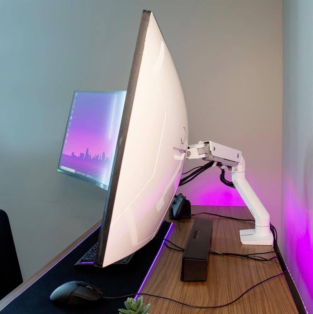 Moderní počítačový monitor na nastavitelném rameni, osvětlený růžovým a modrým světlem, stojící na dřevěném psacím stole.