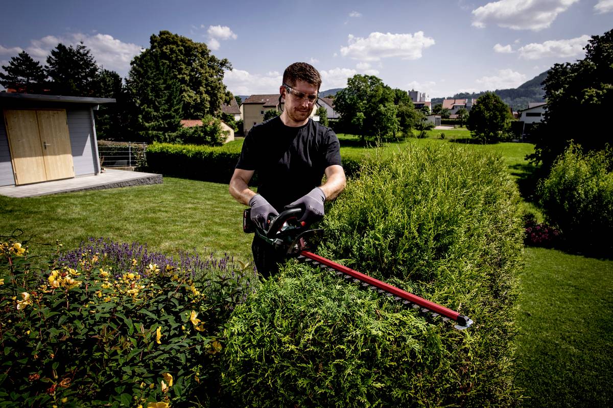 Muž stříhá živý plot elektrickými zahradními nůžkami v zahradě. V pozadí jsou domy a modrá obloha s mraky.