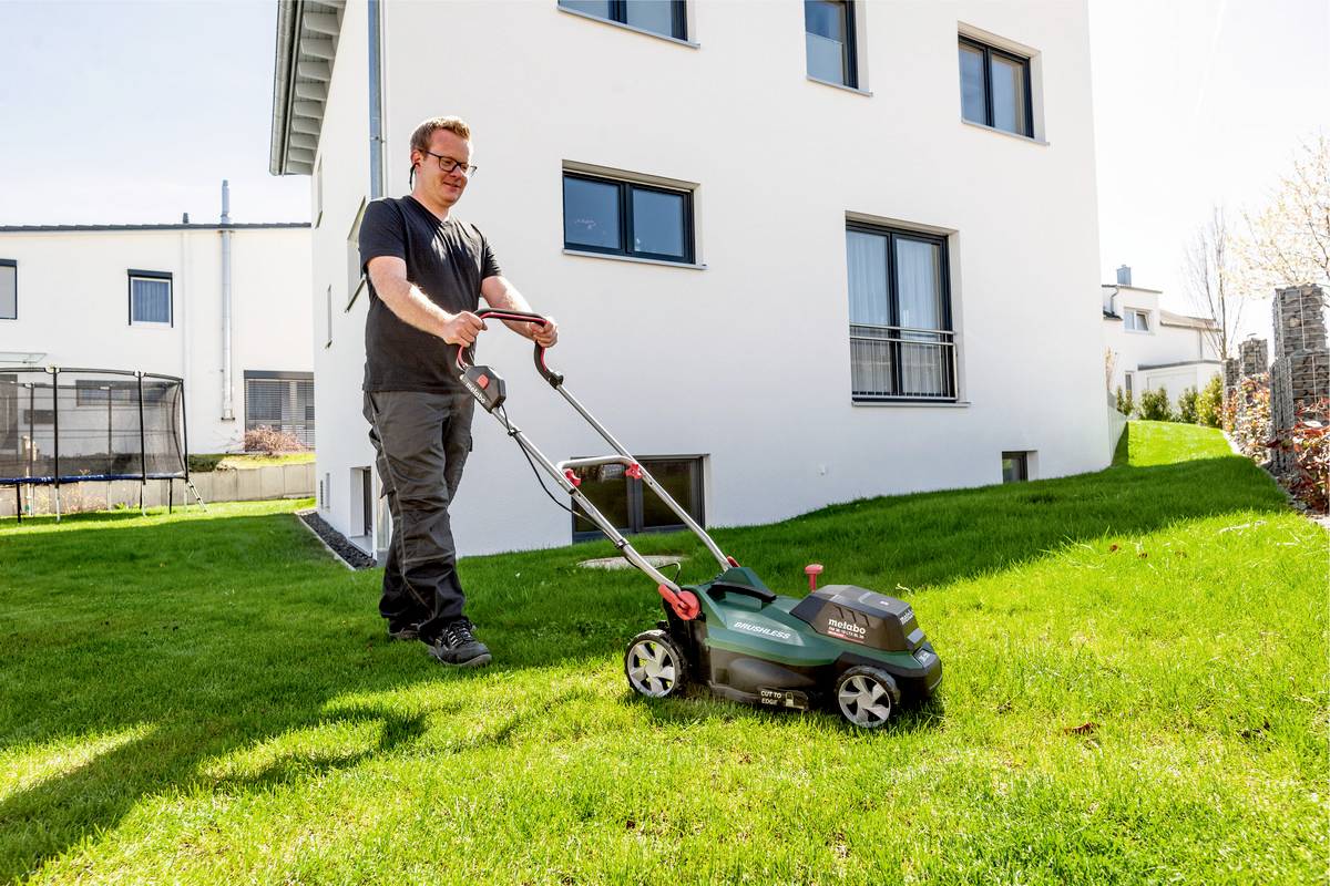 Muž seká trávu elektrickým sekacím strojem před moderním bílým rodinným domem za slunečného počasí.