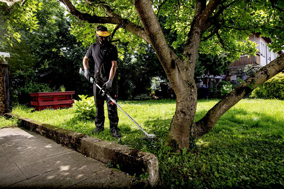 Člověk seká trávu křovinořezem pod stromem v slunné zahradě. V pozadí je vidět červenou lavičku.