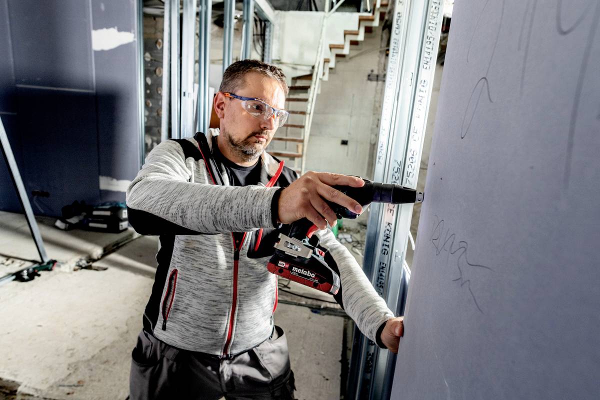 Jeden člověk pracuje na staveništi a upevňuje zeď pomocí akumulátorového šroubováku. V pozadí jsou lešení.