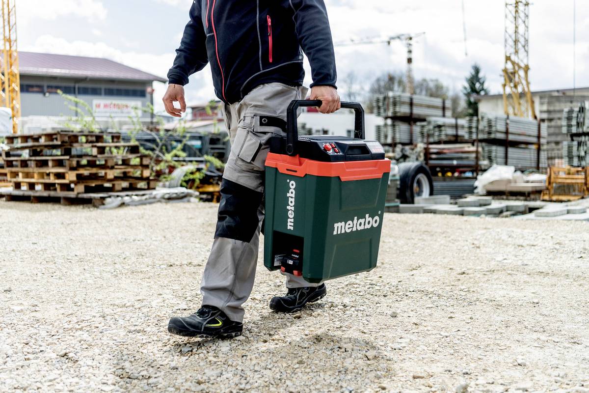 Muž nese zelený kufřík na nářadí značky Metabo na staveništi. V pozadí jsou stavební materiály a jeřáby.