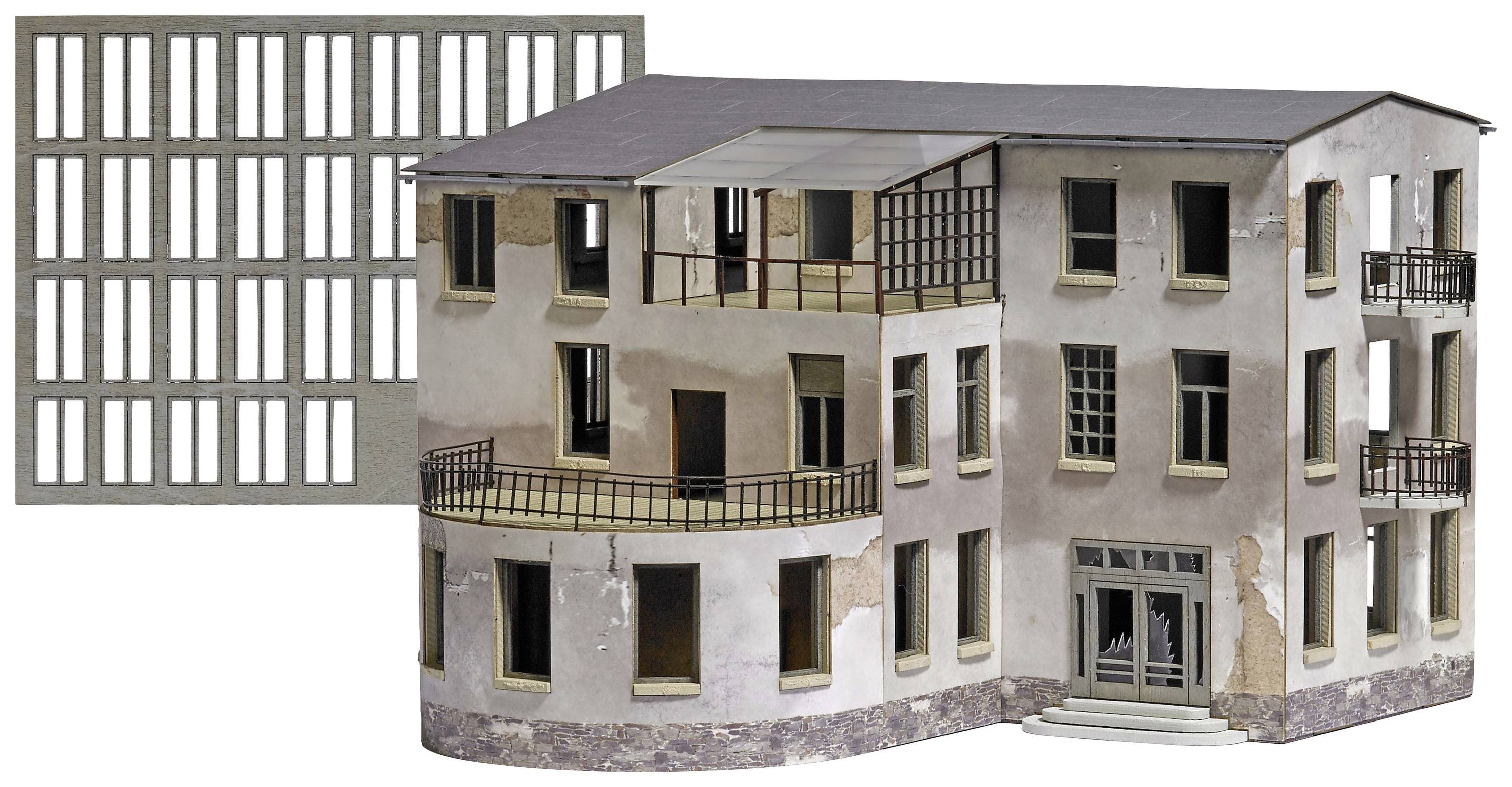 Zchátralá třípodlažní modelová budova s rozbitými okny a balkóny, vedle ní mříž s obdélníkovými otvory.