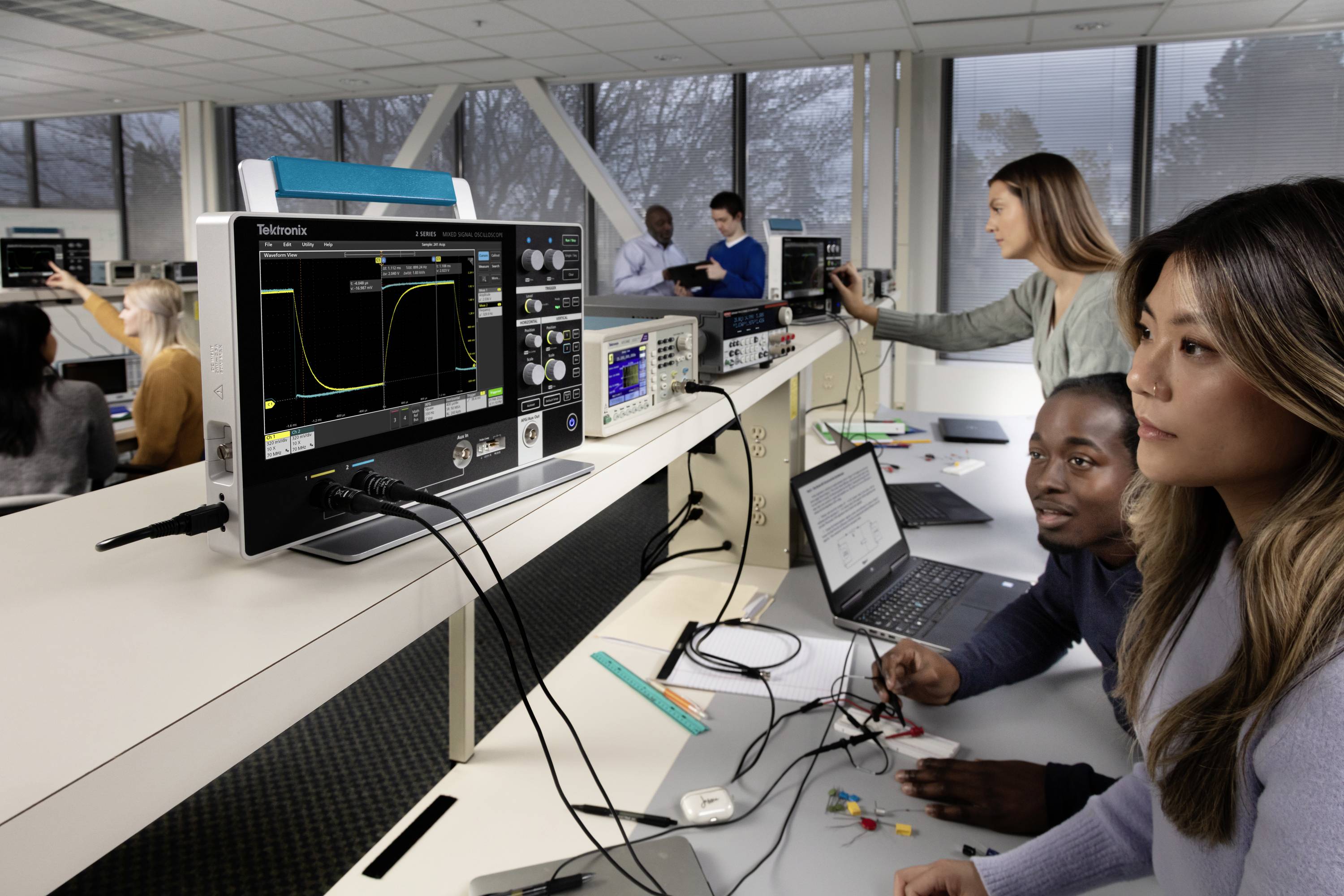 Studenti pracují v laboratoři na elektronických zařízeních a počítačích. Osciloskop zobrazuje průběhy vln.
