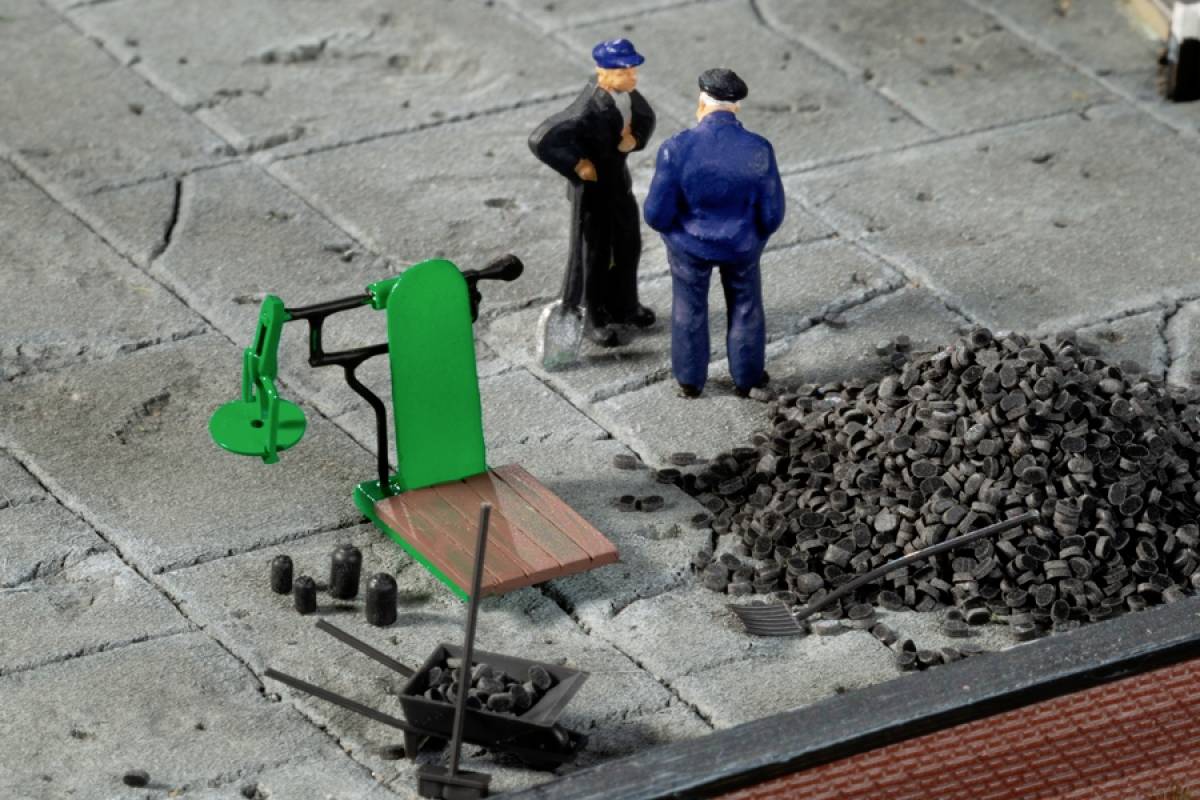 Dvě modelové figurky stojí na dlažbě vedle zeleného zařízení a hromady kamení. Zdá se, že spolu rozmlouvají.