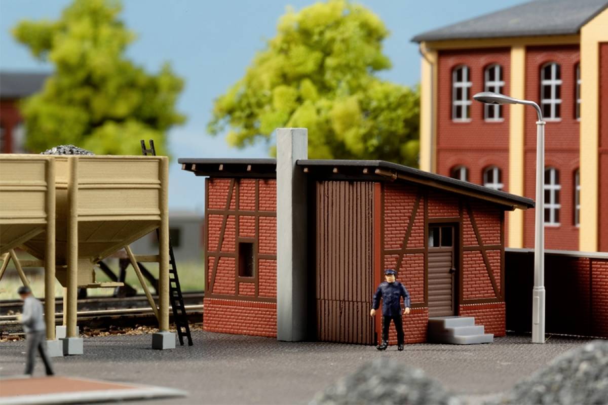 Miniaturní model ukazuje nádražní budovu s hrázděnou architekturou, dvě postavy a budovu v pozadí při modrém nebi.