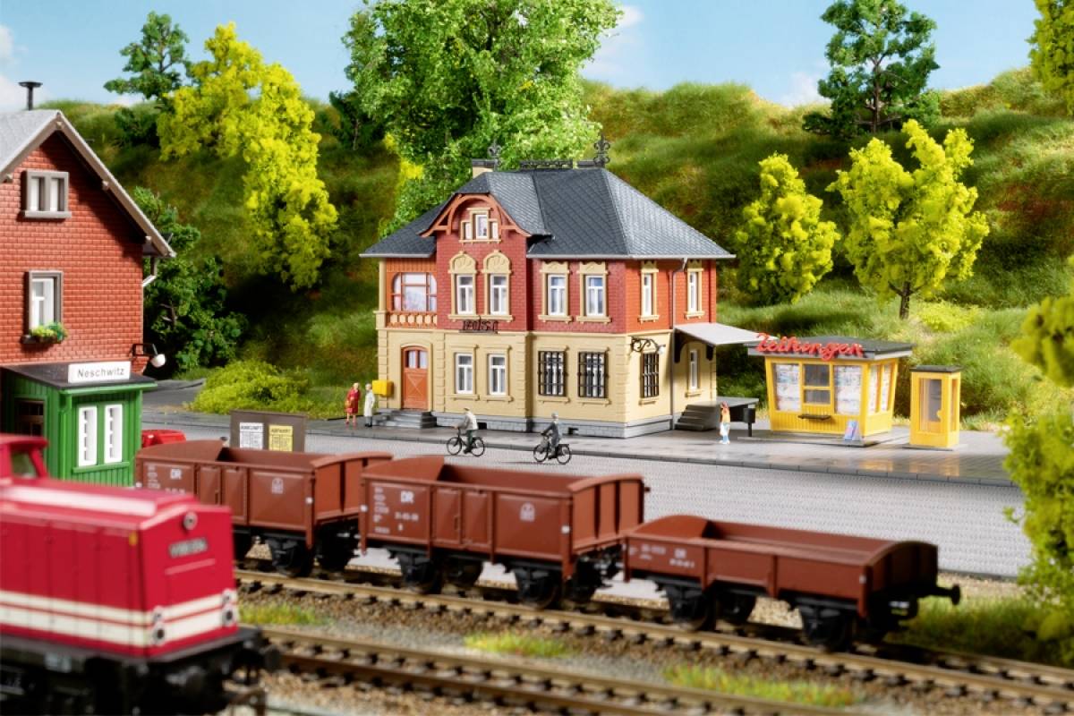 Model železniční scéna s nádražím, nákladními vlaky a budovami v venkovské krajině. Dva osobní jízdní kola stojí vedle nádraží.