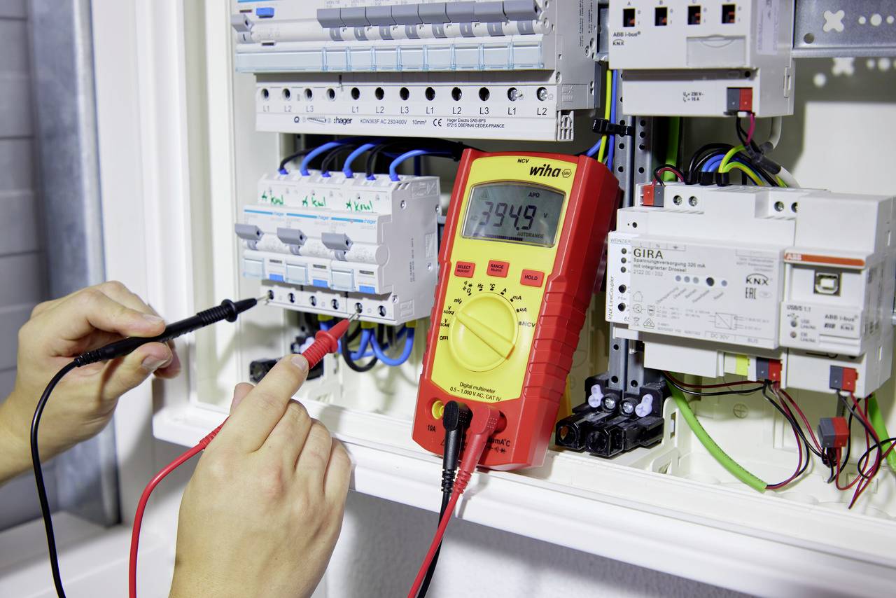 Dvě ruce používají multimetr na otevřené rozvodné skříni k testování elektrických připojení se zaměřením na naměřenou hodnotu 394,9.
