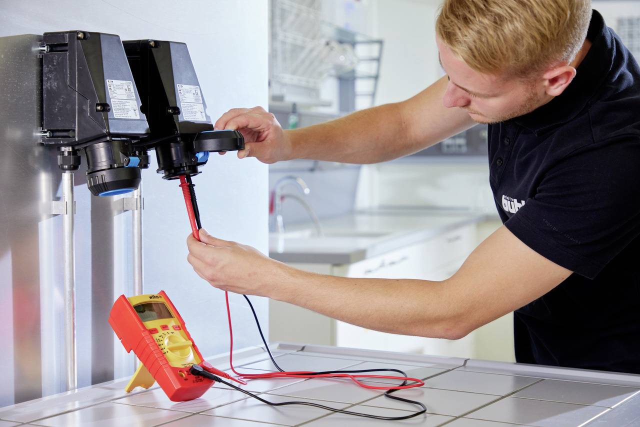 Muž měří elektrické hodnoty multimetrem na průmyslové strojní součásti v prostředí dílny.