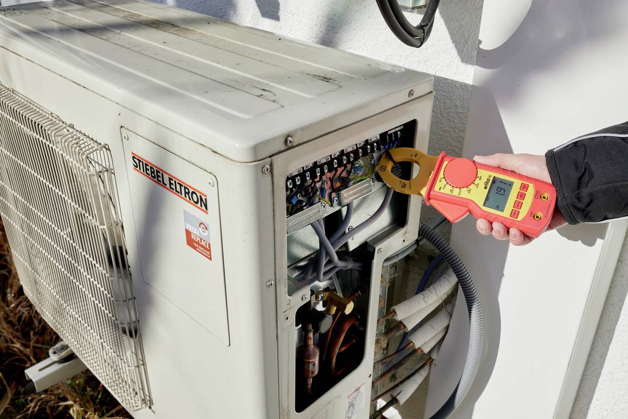 Osoba měří elektrický proud digitálním měřicím přístrojem u jednotky klimatizace za účelem ověření její funkčnosti.