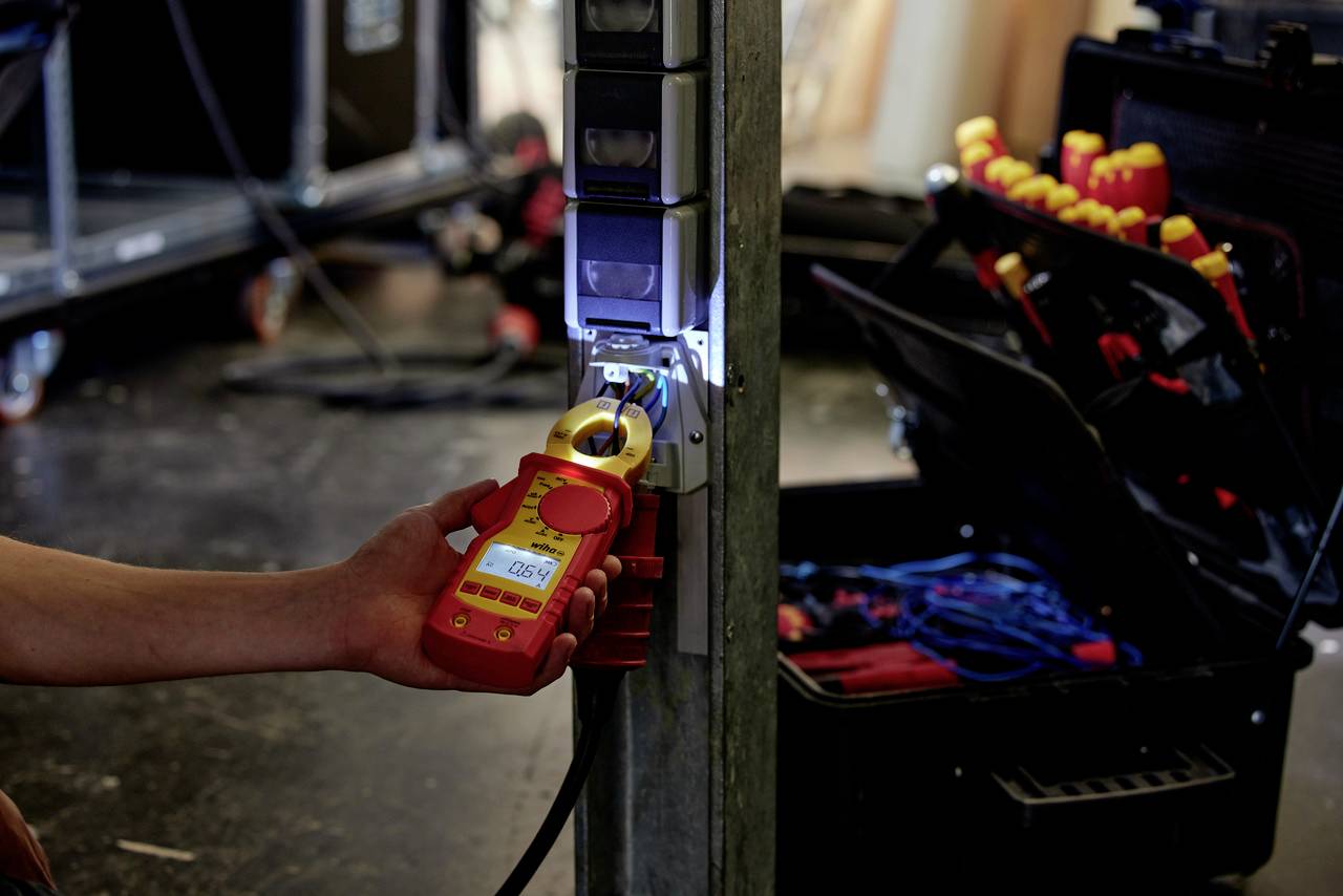 Osoba měří elektrický proud v kabelu digitálním multimetrem. V pozadí leží kufřík s nástroji a šroubováky.