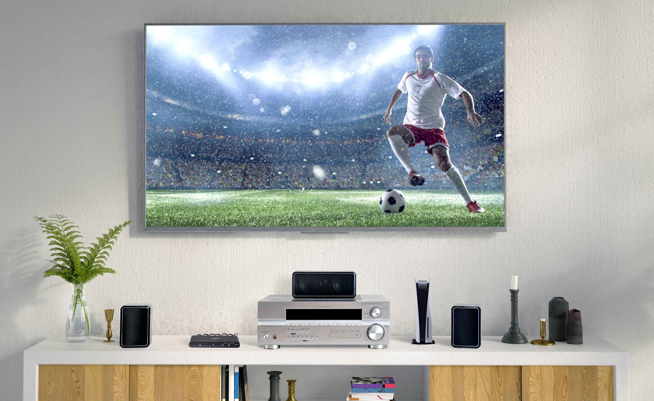 Obývací pokoj s plochým televizorem na stěně, na kterém je vidět fotbalista hrající na stadionovém hřišti. Pod ním stojí police s elektronickými zařízeními a reproduktory.