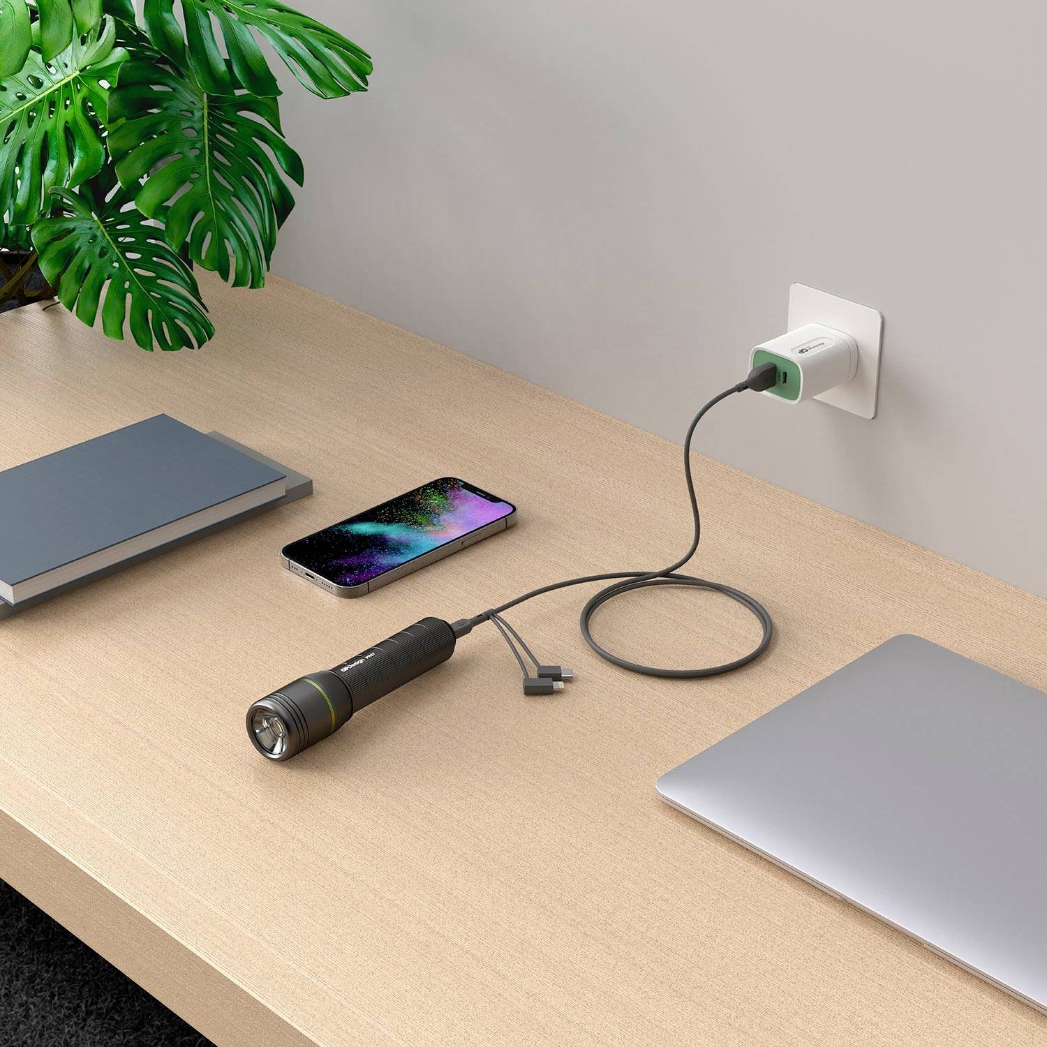 Na stole leží baterka, smartphone, poznámkový blok a laptop. Mobilní telefon je připojen k nabíječce v elektrické zásuvce.