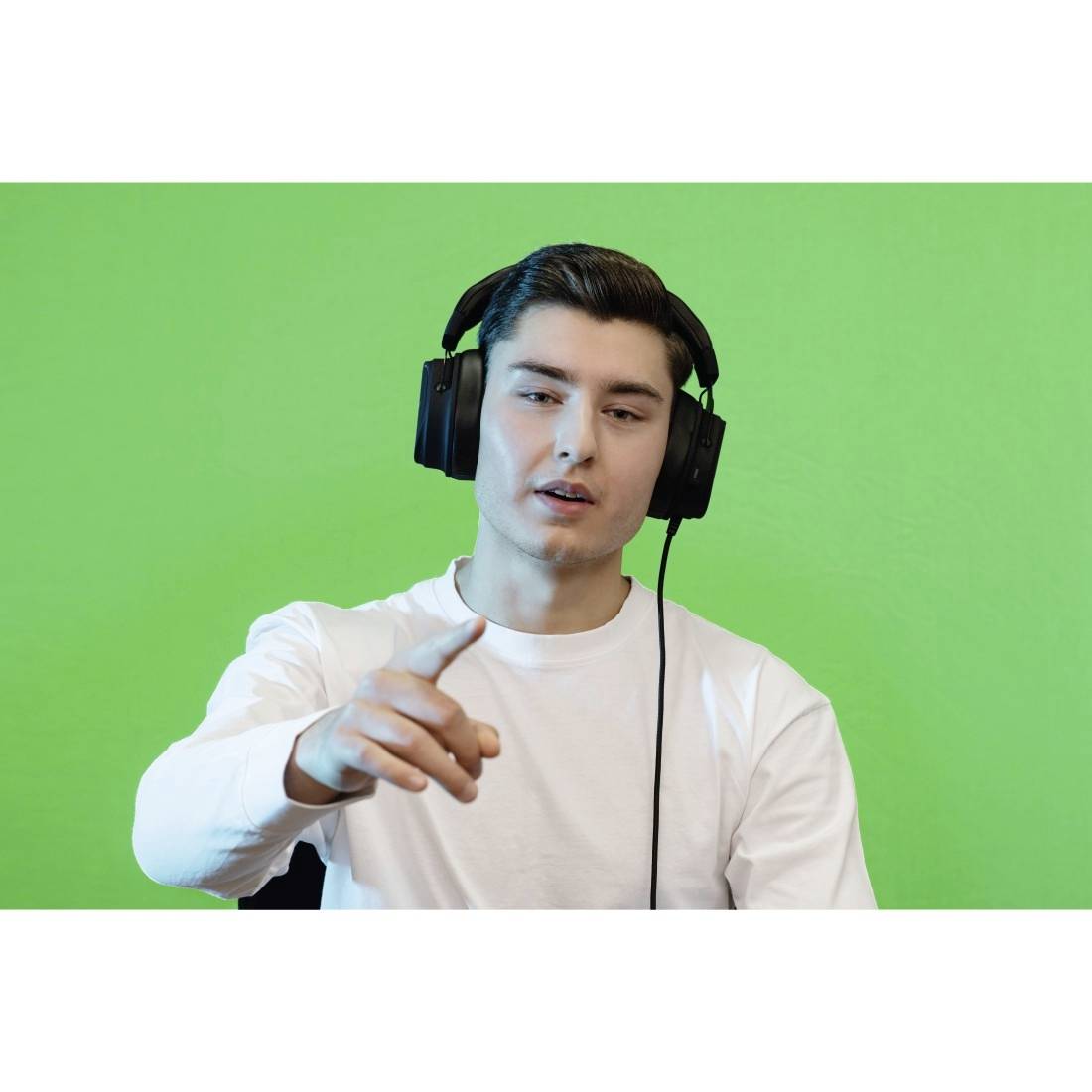 Muž se sluchátky a bílým tričkem sedí před zeleným pozadím a ukazuje dopředu.