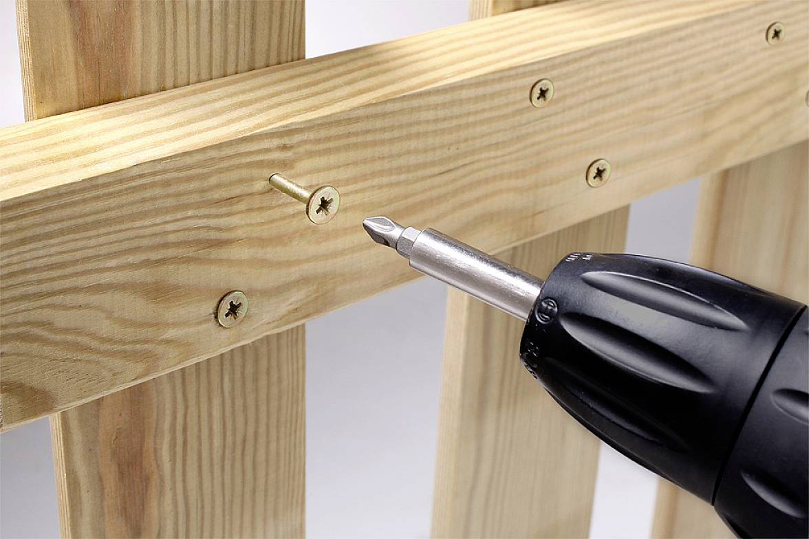 Akumulátorový šroubovák zatahuje šroub do dřevěné latě na paletě, několik šroub již je upevněno.