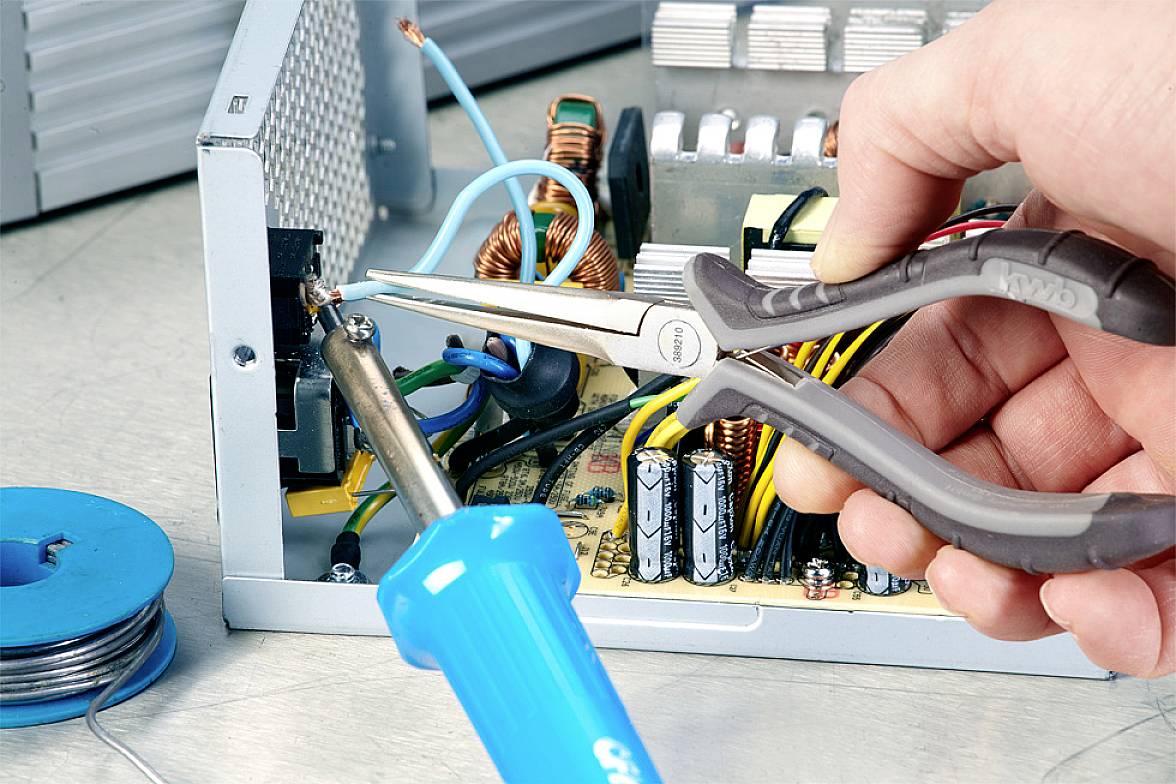 Jedna ruka drží kleště při opravě elektronického zařízení. Pájecí pero a dráty jsou viditelné.