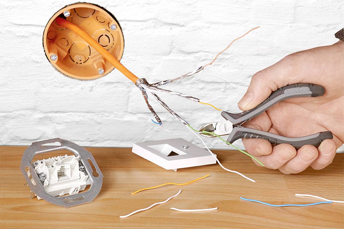 Jedna ruka drží kleště a odstraňuje izolaci z kabelů, které vyčnívají z oranžové krabice ve zdi.
