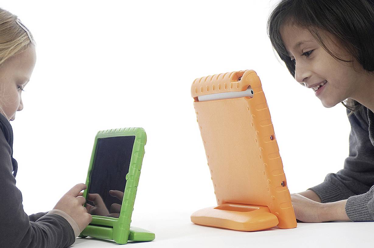 Dvě děti sedící naproti sobě si každé hrají s tabletem. Dívka vlevo používá zelený tablet, dívka vpravo má oranžový tablet.