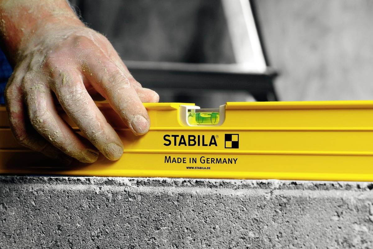 Jedna ruka drží žlutou vodní váhu s nápisem 'STABILA Made in Germany' na šedém kamenném bloku.