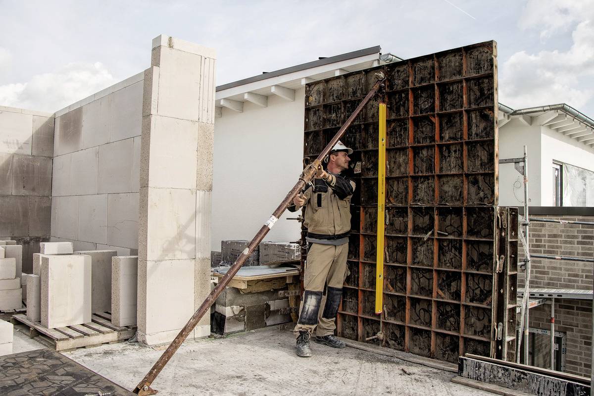 Dělník staví zeď na staveništi. Má na sobě ochranný oděv a používá opěru k zajištění zdi. V pozadí jsou vidět další budovy.