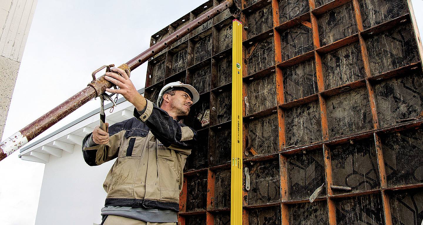 Dělník v přilbě a s nástroji staví bednění na staveništi. V pozadí jsou viditelné lešení.