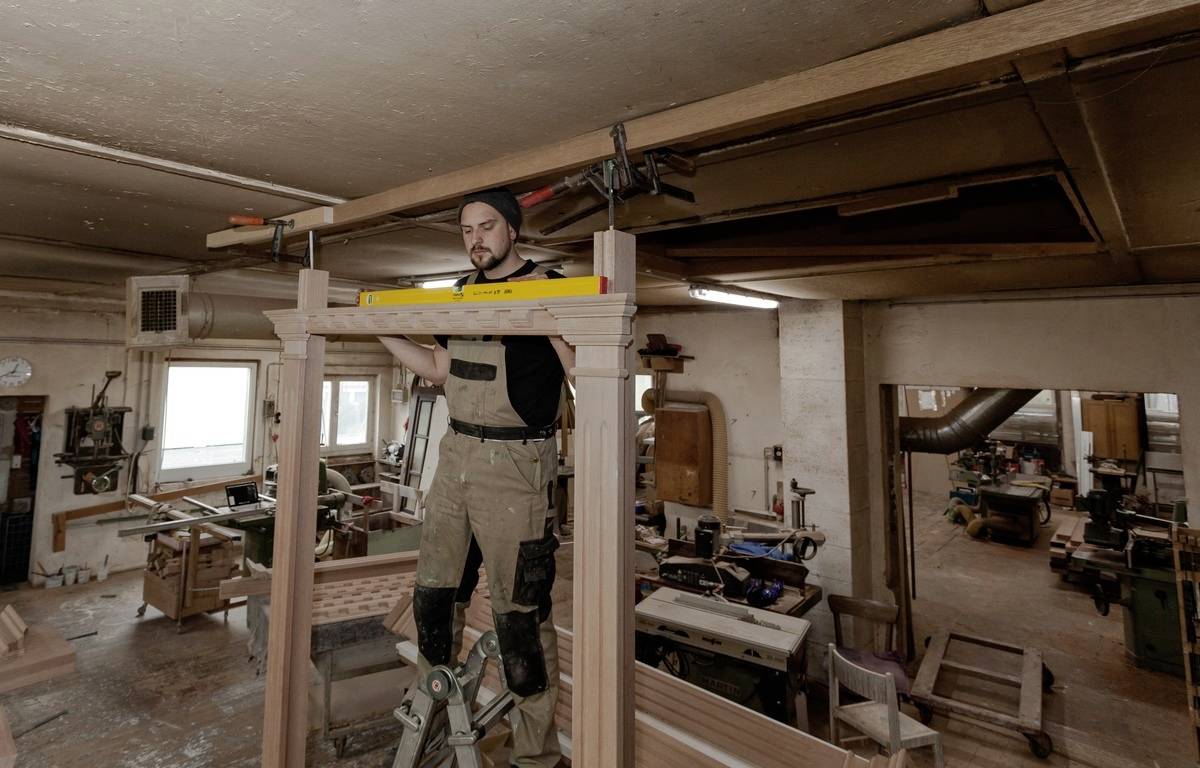 Řemeslník stojí na žebříku v dobře vybavené dílně a pomocí vodováhy kontroluje horizontální rovinu dřevěné konstrukce.