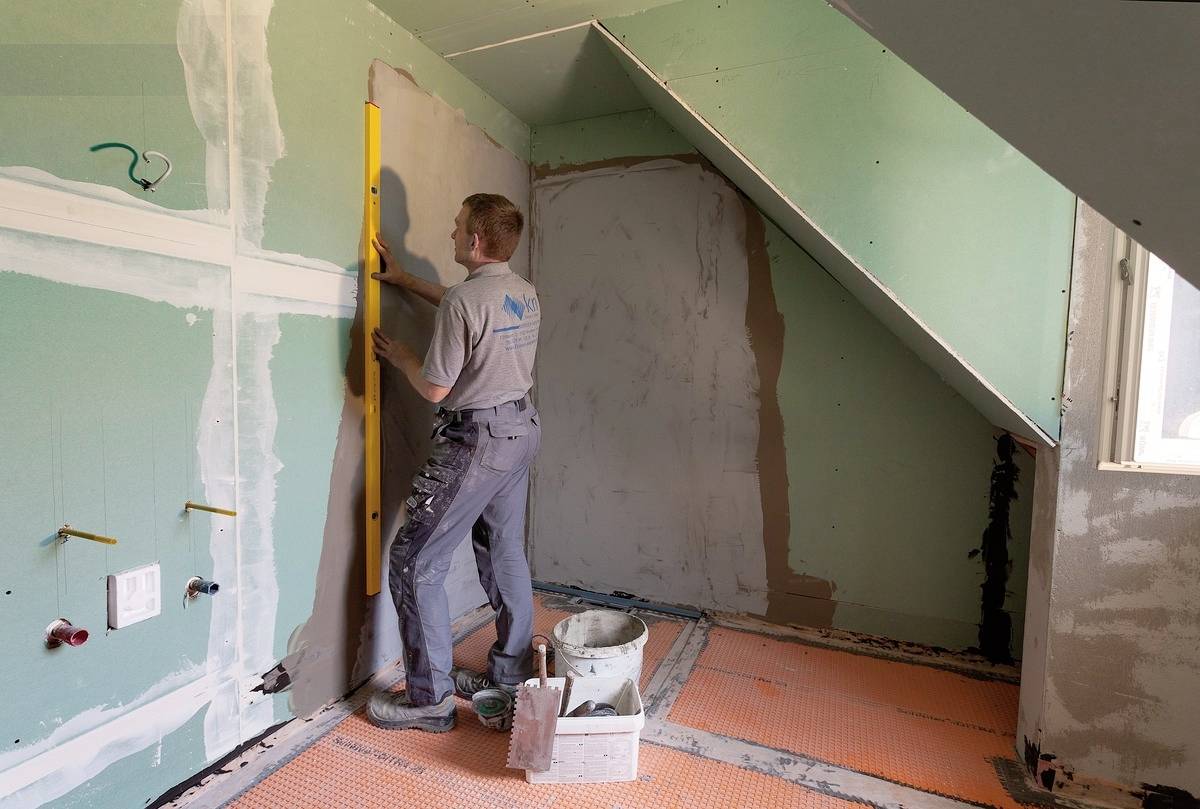 Muž na staveništi vyrovnává nástěnné desky pomocí vodováhy. Místnost má šikmé stěny a je částečně neomítnutá.