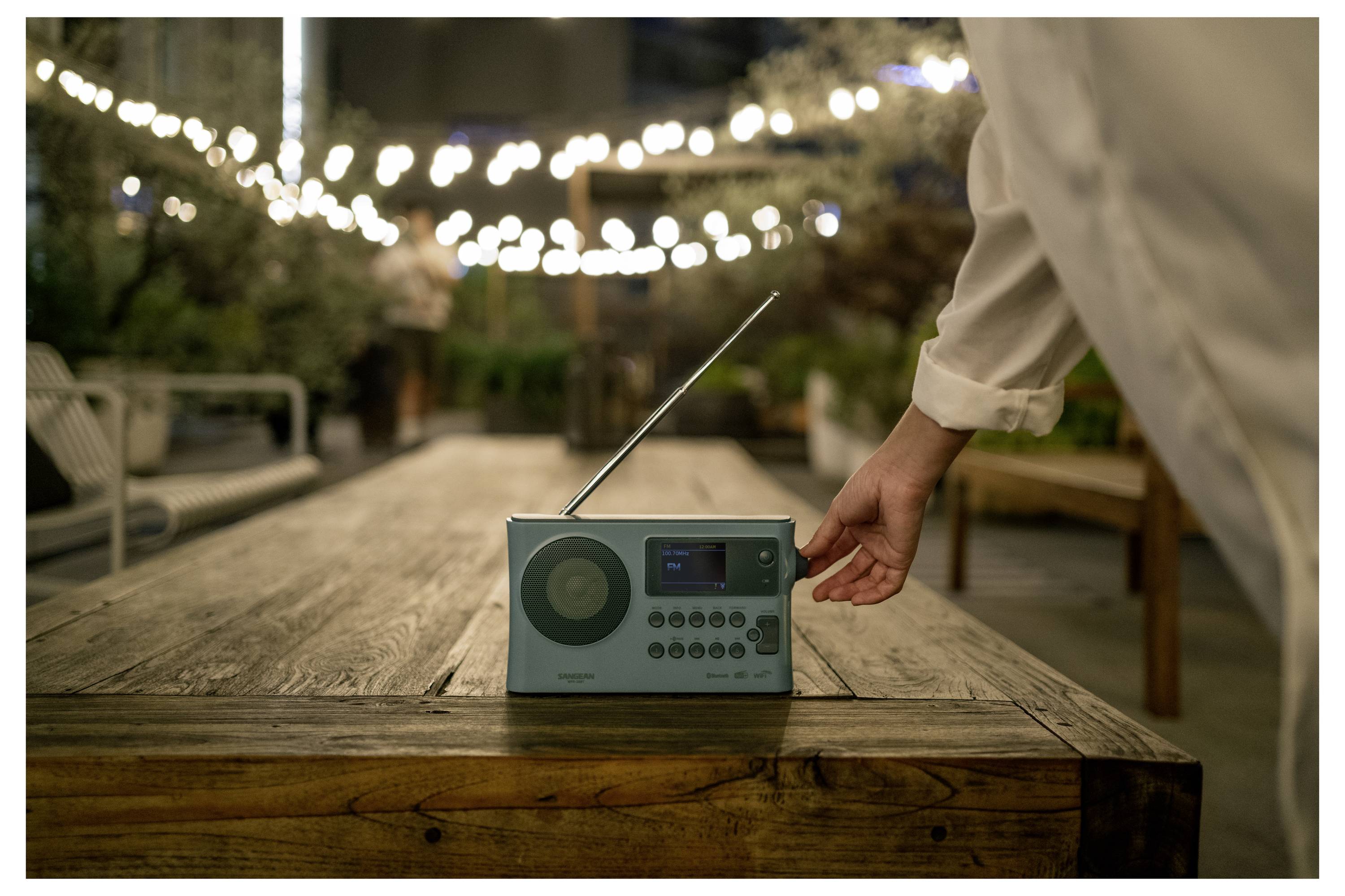 Ruka pokládá tyrkysové přenosné rádio na dřevěný stůl venku pod řetězy světel v soumraku.