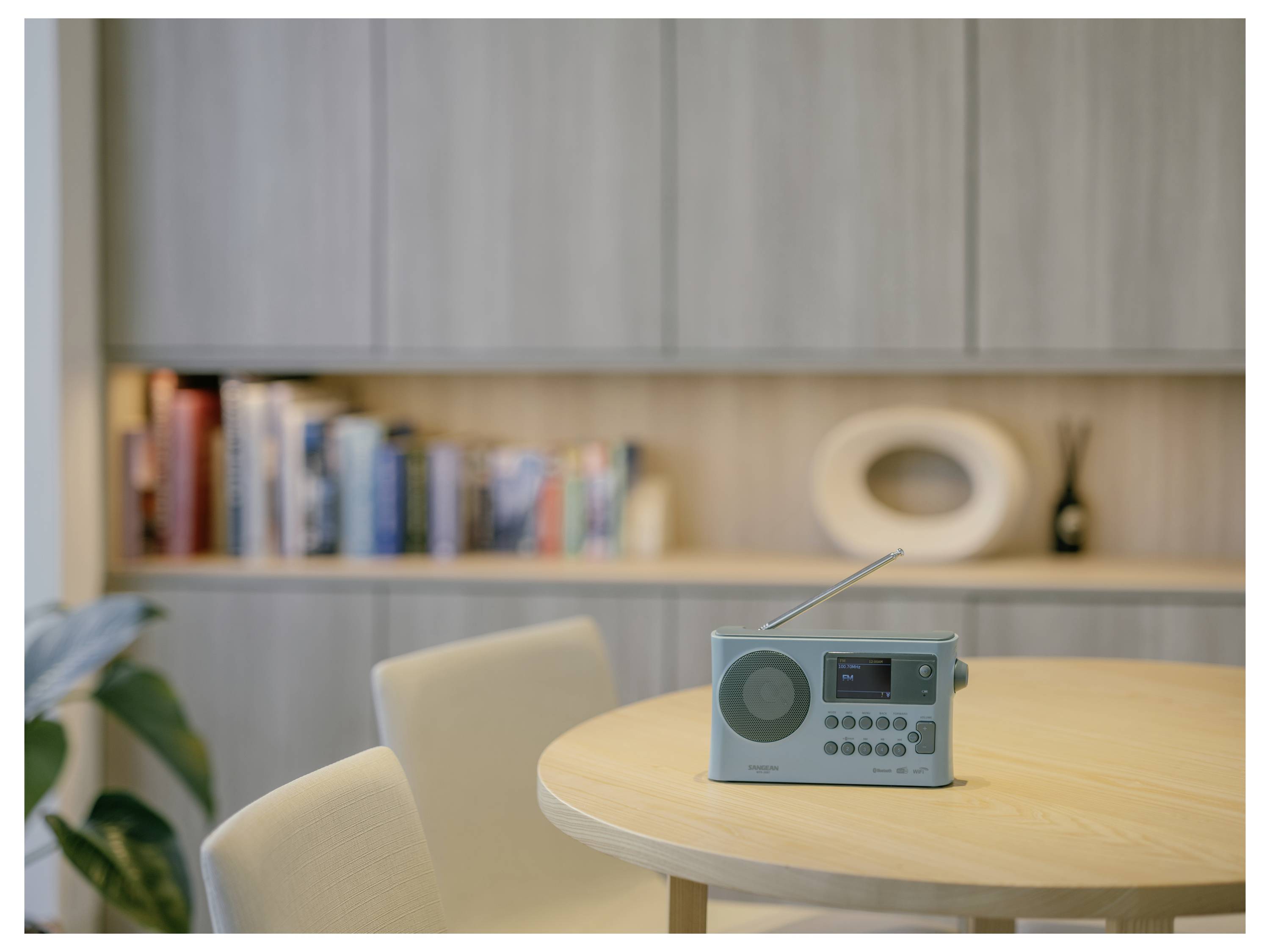 Malý přenosný rádio sedí na kulatém stole v moderní místnosti s policemi a skříňkami na pozadí.