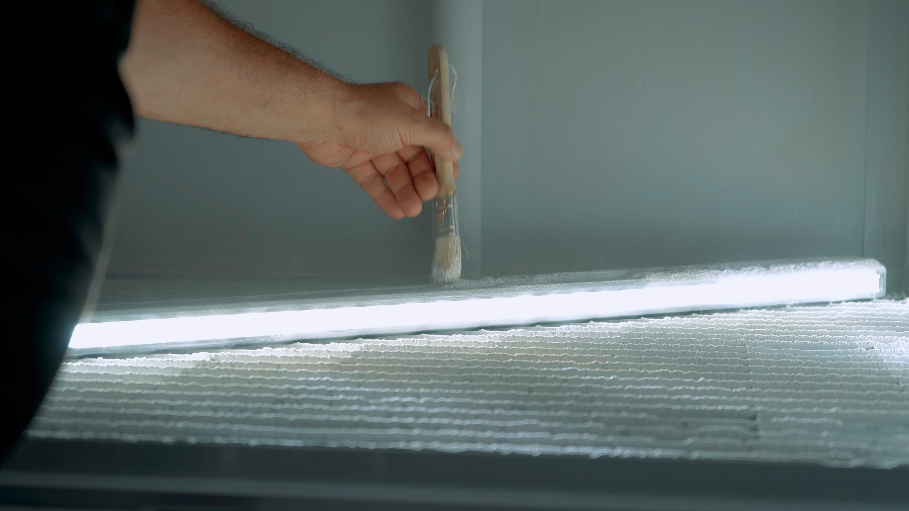 Osoba štětcem natírá svítící LED lampu upevněnou na texturovaném povrchu.