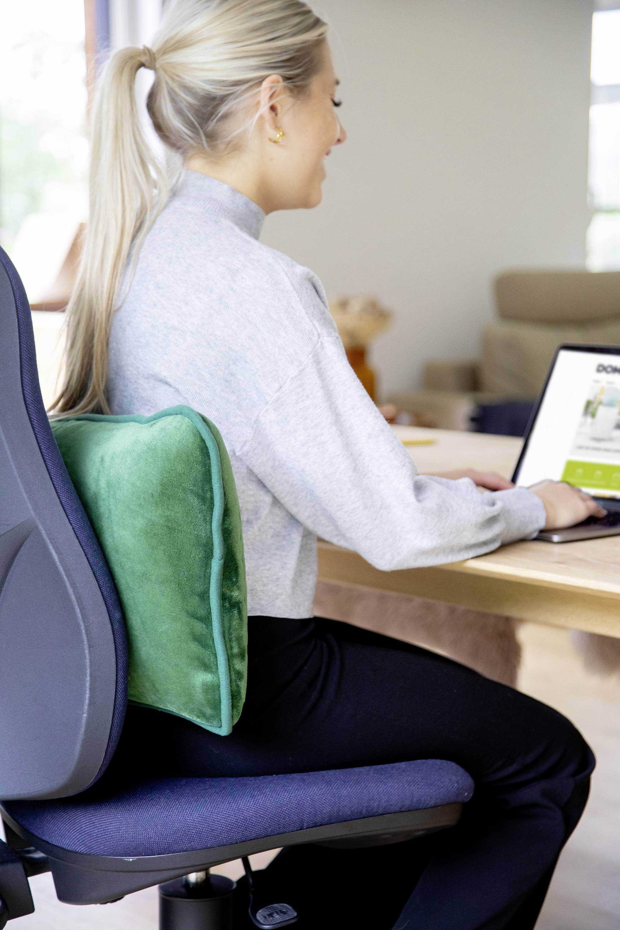 Žena sedí na kancelářské židli a pracuje na laptopu. Zelený polštář jí podporuje záda.
