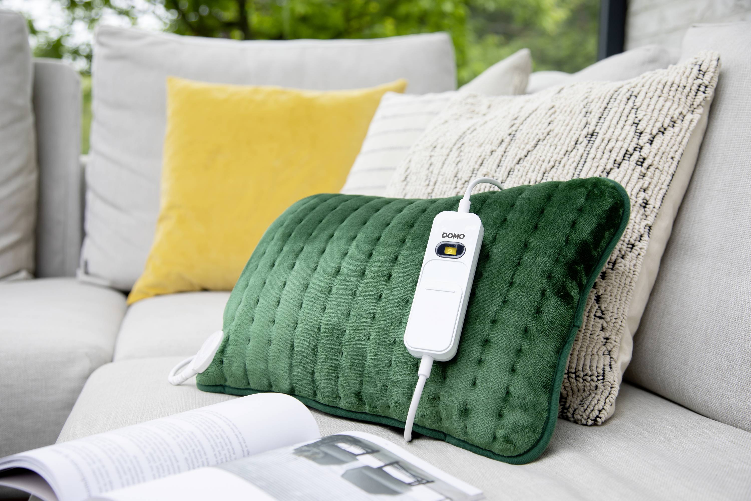 Zelený elektrický nahřívací polštář s kabelovým ovládáním leží na gauči s žlutým a béžovým vzorovaným polštářem v pozadí.