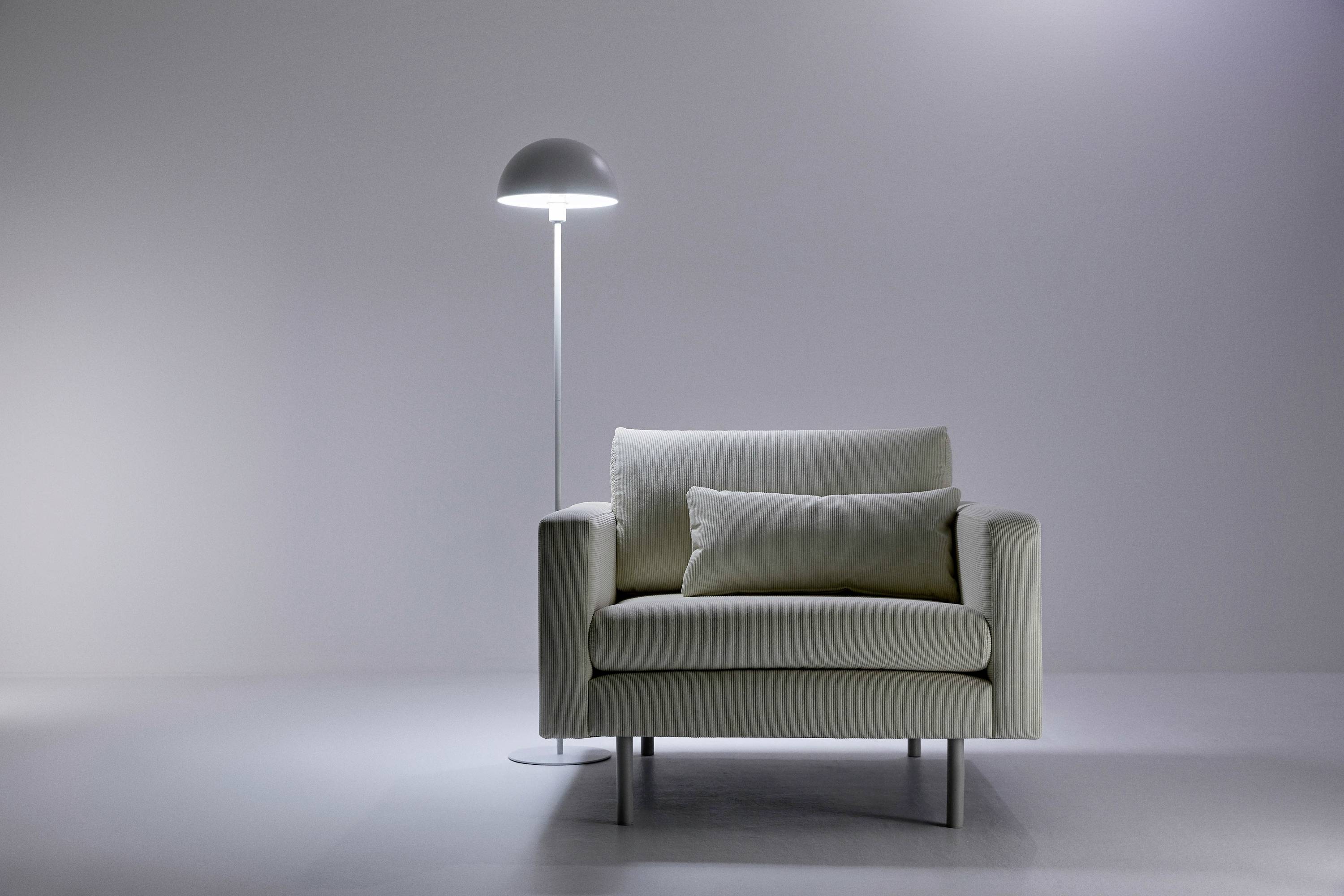 Moderní, krémový křeslo stojí na světlé podlaze, vedle štíhlé bílé stojací lampy. Minimalistický design.