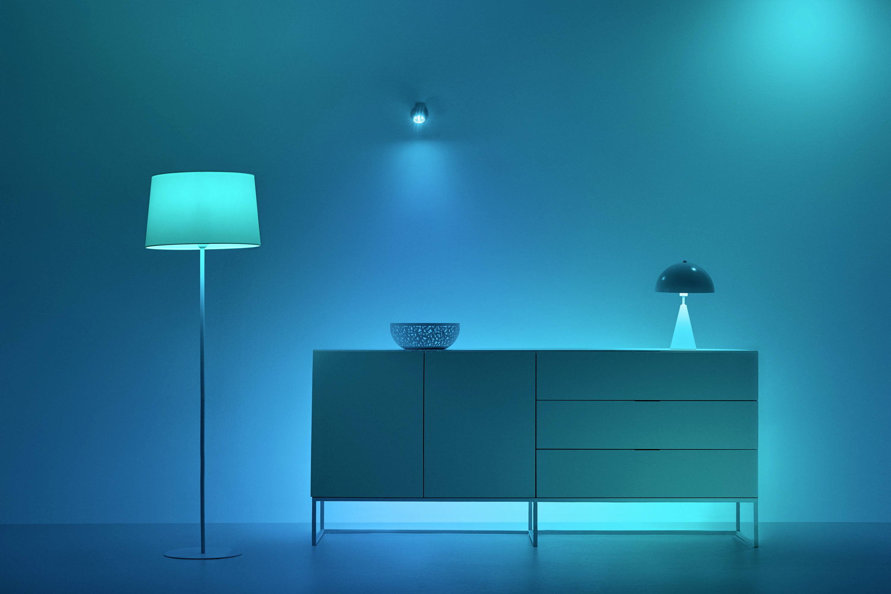 Minimalistický interiér obývacího pokoje s modrým osvětlením, který zobrazuje stojací lampu, příborník s mísou a stolní lampou.
