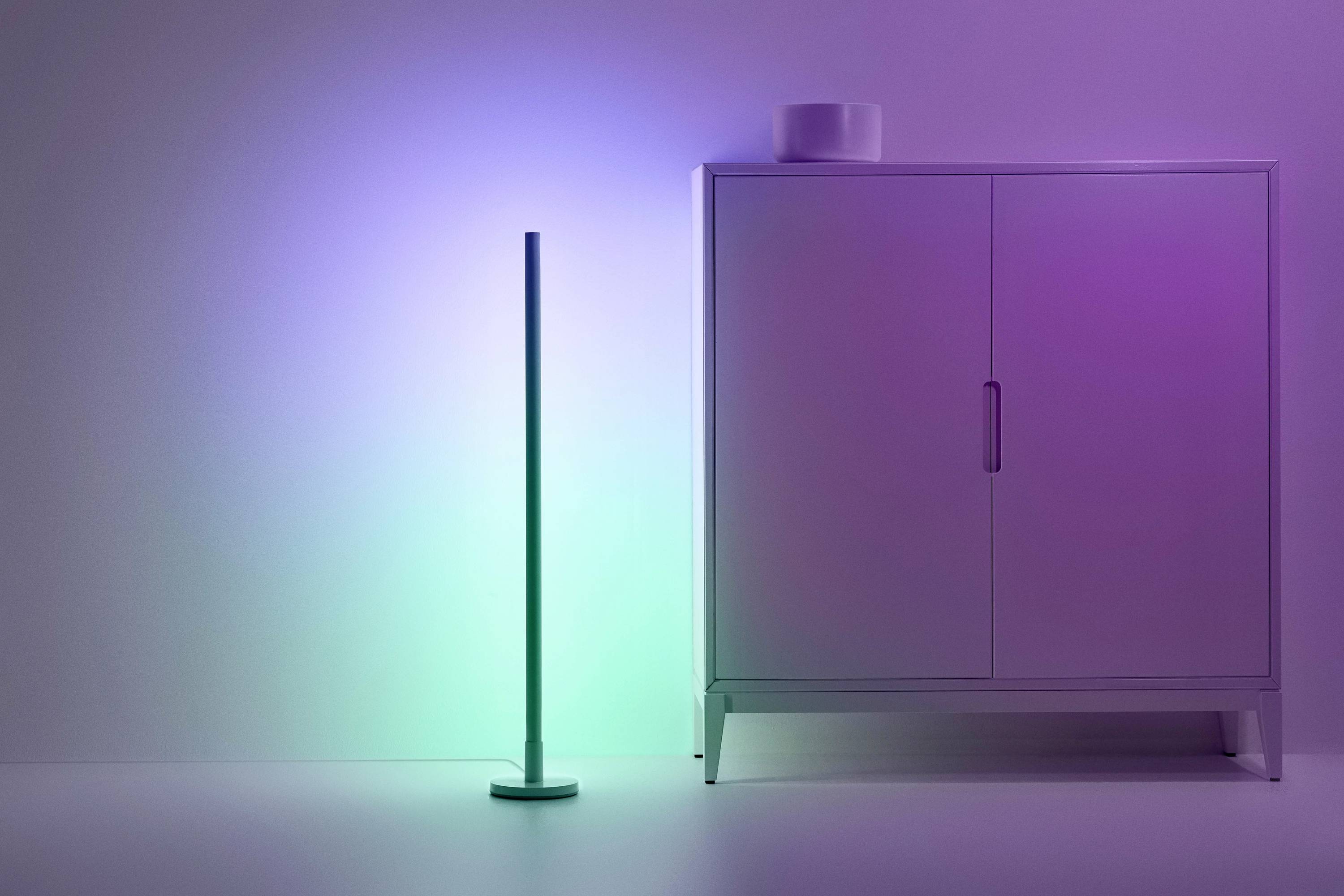 Stojací lampa svítí v pestrých barvách vedle bílé skříně před bílou stěnou. Ambientní osvětlení v moderním prostoru.