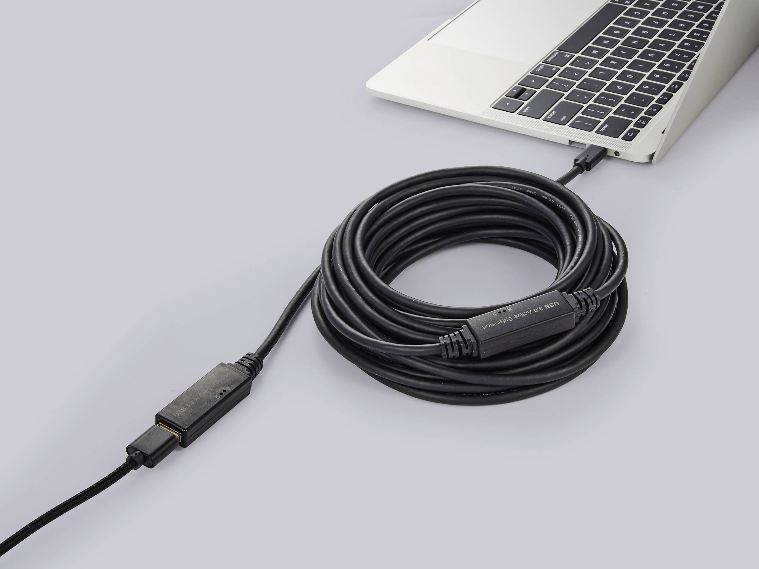 Černý HDMI kabel je připojen k otevřenému laptopu. Kabel je smotán a uprostřed vybaven zesilovačem.